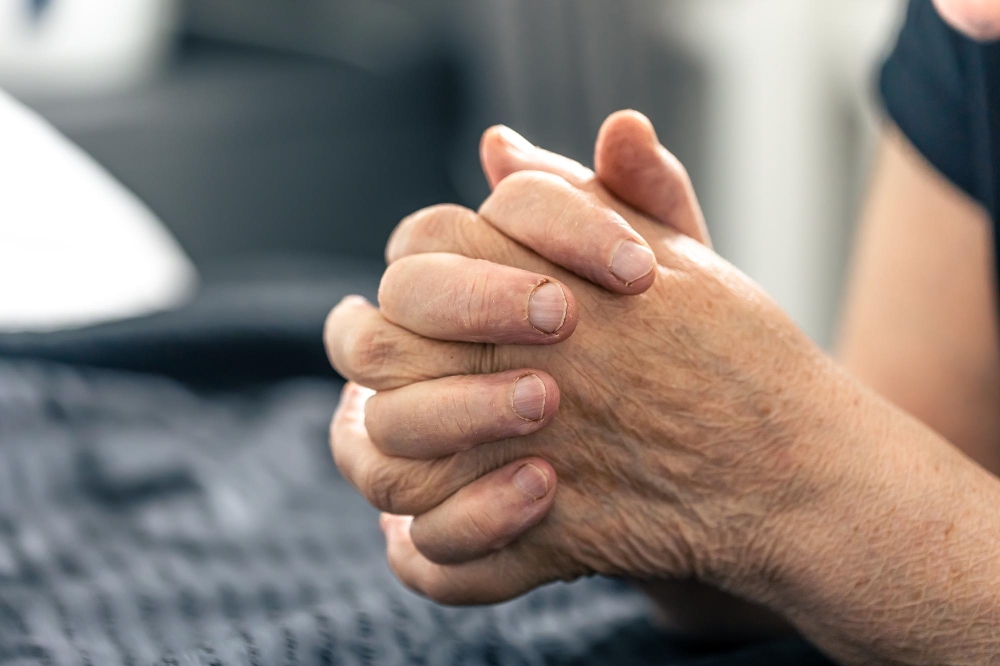 Elderly hands clasped together in close-up, resting on dark textured blanket.